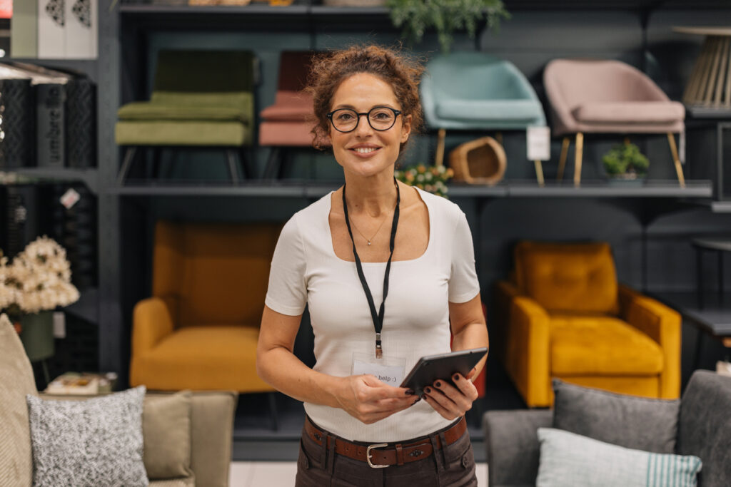 A smiling woman stands in a modern furniture store, holding a tablet while surrounded by colorful seating options, demonstrating her enthusiasm and engagement in retail.